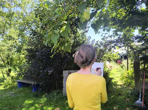 Eine Zeichnerin steht mit dem Rücken zum Betrachter, zur Betrachterin im Garten und zeichnet Freihand eine Auftragszeichnung. Sie hat ein gelbes Oberteil an, im Garten steht eine Bank im Schatten.